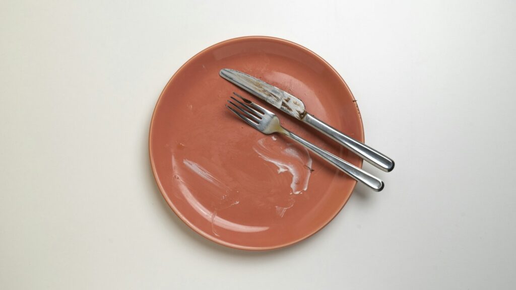 An empty coral-coloured plate with a fork and knife laid across it on a white table