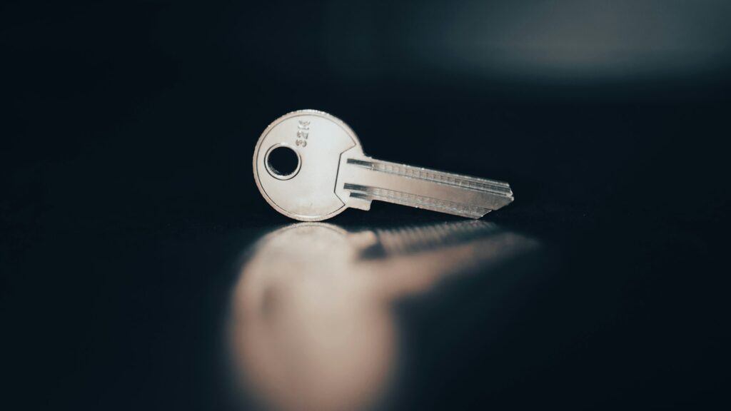 A single silver key lying on a dark surface, reflected in its glossy finish