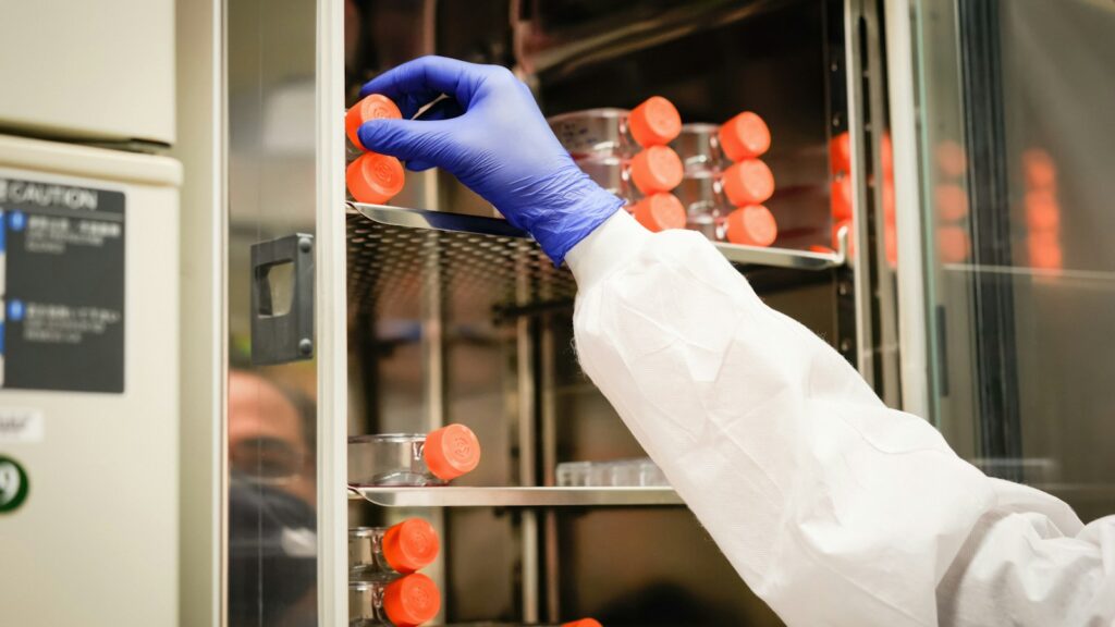 A researcher in a white coat and blue gloves reaching for cell cultures in a laboratory incubator